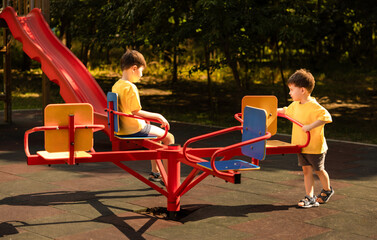 Happy children playing outside in a park on a roundabout with seats. Siblings enjoy a sunny summer day, spinning together and smiling. Kids wearing yellow tank tops having fun outdoors.