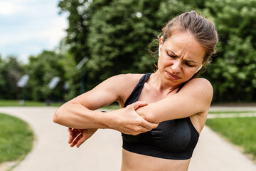 Female runner standing and holding painful elbow with pained expression during outdoor workout