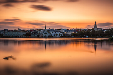 A serene sunset over Reykjav&iacute;k reflecting on the still waters of Tj&ouml;rnin lake, with iconic landmarks like Hallgr&iacute;mskirkja and Fr&iacute;kirkjan silhouetted against the glowing sky