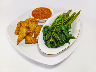 Meal on a white square plate with a gold rim, consisting of fried tempe, boiled cassava leaves, and a small bowl of sambal. Related to Indonesian cuisine, local meals, traditional home cooking