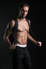 Fototapeta premium A muscular man stands confidently against a dark backdrop, showcasing his toned physique while holding a backpack strap. The emphasis is on fitness and strength in a well-lit studio