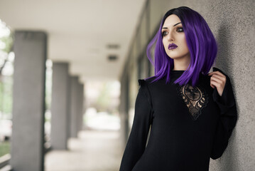 Obraz premium A gothic woman with striking purple hair stands against a concrete pillar, showcasing a confident demeanor. She wears a sleek, dark outfit, blending modern fashion with an edgy style