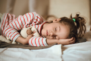 A young girl in striped pajamas sleeps soundly in a bright bedroom, cradling a teddy bear. Soft light illuminates her serene expression while she rests on a comfortable bed