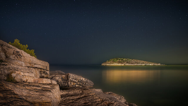 Starry night over a tranquil sea with a rocky coastline and a distant island