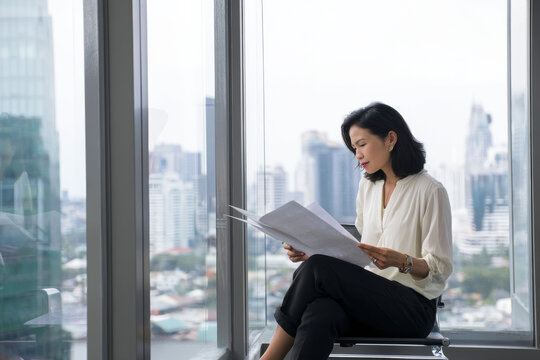 Middle-aged Asian business woman sitting in her office, reading through reports while preparing for a presentation, dressed in a formal blouse and black trousers, the office clean and professional