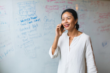 Middle-aged Asian businesswoman on a coaching call, whiteboard filled with goals behind her, giving career advice and empowerment strategies with clarity and warmth