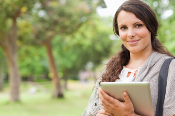 Female wearing gray cardigan over white blouse holding tablet and bag in leafy park, copy space