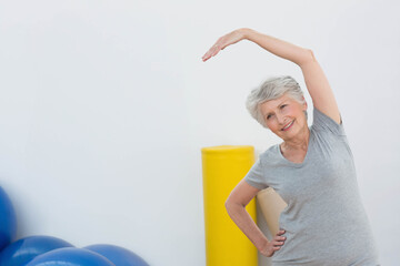 Senior woman performing side stretch at fitness studio with foam roller and balls, copy space