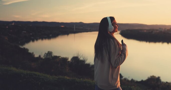 Young woman wearing headphones listening to music and dancing by the river at sunset during autumn