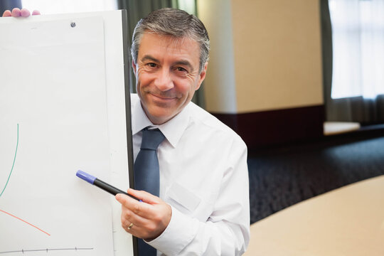 Mature businessman in shirt and tie standing by flip chart explaining line graph in conference room