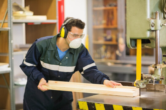 Woodworker operating vertical bandsaw cutting long wooden plank in workshop near metal shelving