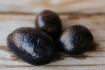 Close-up view of a whole raw purple potato with dark skin placed on a wooden surface in soft natural light. Shallow depth of field and visible texture.