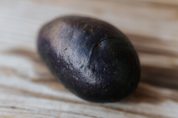 Close-up view of a whole raw purple potato with dark skin placed on a wooden surface in soft natural light. Shallow depth of field and visible texture.