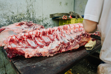 A butcher cleans a large rack of raw beef ribs on a wooden cutting board in a dimly lit, industrial setting.