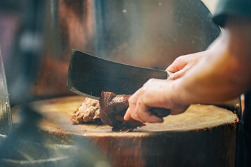 A person's hand chops meat on a wooden cutting board with a large cleaver. Smoke rises from a pot in the background.