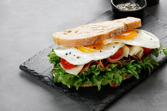 Sandwich, toast with eggs, bacon, cheese, green lettuce on black slate board. Fresh and healthy breakfast.