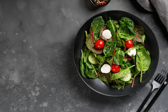 Black plate of Fresh green salad with arugula, spinach, cucumber, cherry tomatoes, mozzarella.