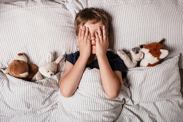 The boy was scared before going to bed. Night terrors in a child. The kid covers his face with his hands in a fear. Children's experiences. Boy in the bed. View from above. Sad psychological state.