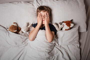The boy was scared before going to bed. Night terrors in a child. The kid covers his face with his hands in a fear. Children's experiences. Boy in the bed. View from above. Sad psychological state.