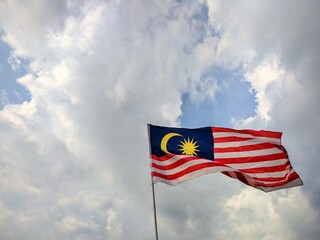 The national flag of Malaysia, known as the Jalur Gemilang, is seen waving prominently against a dramatic cloudy sky. 