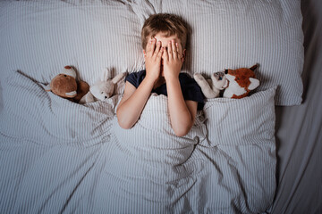 The boy was scared before going to bed. Night terrors in a child. The kid covers his face with his hands in a fear. Children's experiences. Boy in the bed. View from above. Sad psychological state.