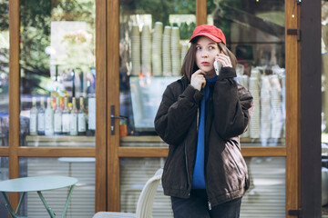 Fototapeta premium Young woman in hoodie and cap talking on smartphone outside a cafe.