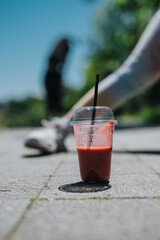 Close-up of a red smoothie cup on pavement with blurred background, showing a vibrant and refreshing outdoor atmosphere with optional fitness context.