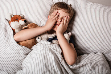 The boy was scared before going to bed. Night terrors in a child. The kid covers his face with his hands in a fear. Children's experiences. Boy in the bed. View from above. Sad psychological state.