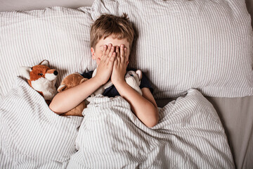 The boy was scared before going to bed. Night terrors in a child. The kid covers his face with his hands in a fear. Children's experiences. Boy in the bed. View from above. Sad psychological state.