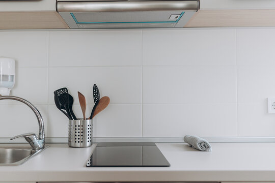 Close-up view of modern stove area with faucet, stainless utensil holder and clean surface. Light tones and simple tools suggest function and tidiness.Modern kitchen with built-in stove, faucet,