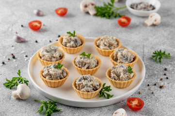 Mini tartlets with mushrooms in a cream sauce, with cheese, garlic and herbs on a white plate on a gray concrete background.