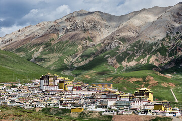 Aerial view of Gongsa Monastery nestled amidst rolling green hills, capped by majestic, rocky mountains under a dramatic sky, Gongsa, Qinghai, China.