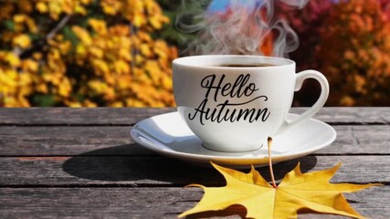 White cup of hot steaming drink with "Hello Autumn" text and yellow maple leaf on a wooden table against blurred autumn park landscape background.Fall season greeting card concept,4K. - Powered by Adobe