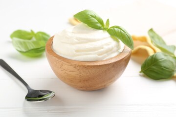 Delicious yogurt in bowl, basil and spoon on white wooden table, closeup