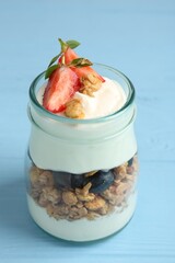 Tasty yogurt with granola and berries in glass jar on light blue wooden table, closeup