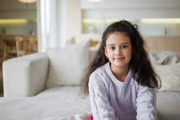 Naklejka premium Smiling girl in cozy sweater enjoys indoor relaxation at a modern living space filled with natural light and minimalist decor