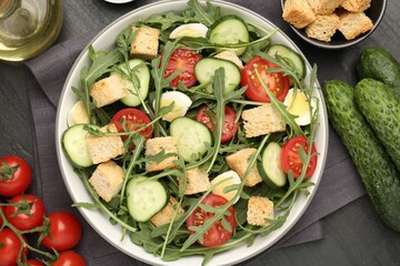 Tasty salad with croutons, cucumbers, tomatoes and arugula on black table, flat lay