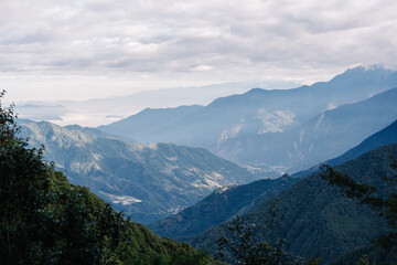 Naklejka premium Panoramic view of a hazy mountain range under a cloudy sky. Lush green vegetation frames the scene.