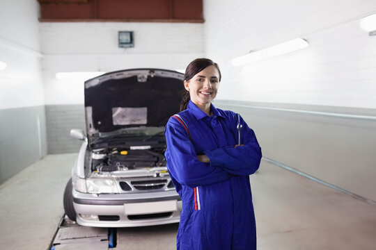 Female mechanic posing in workshop bay in blue coverall near open silver sedan hood showing engine