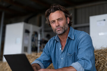 Fototapeta premium A thoughtful man sitting on hay while working on a laptop in a rustic environment, representing the blend of agriculture and modern technology in daily life.