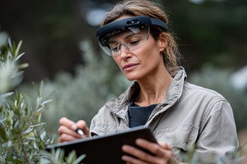 A focused woman in an olive grove utilizes smart glasses to take notes, highlighting the integration of wearable technology in agricultural management and productivity.