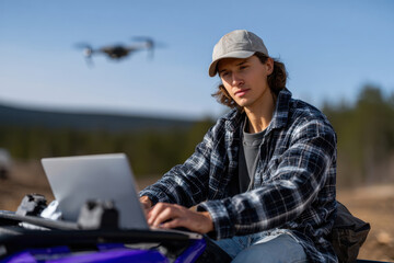 A young man operates a laptop while sitting on an ATV, with a drone flying overhead, exemplifying the integration of technology in outdoor adventure and farming.