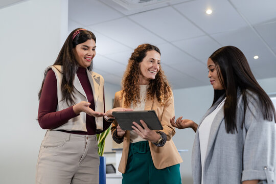 Diverse women colleagues wearing blazers collaborating around digital tablet in office with plant