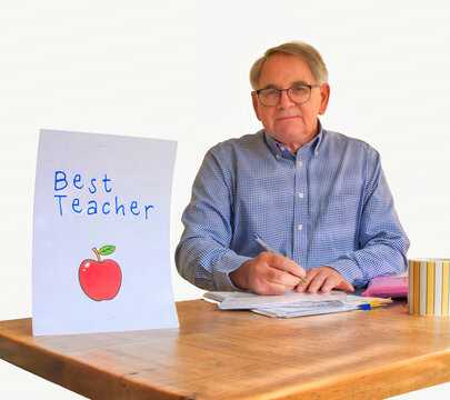 Mature male teacher sitting at his desk mark homework with a Best Teacher card written in a child handwriting an cup sitting in the afternoon sun with sun streaming through the windows