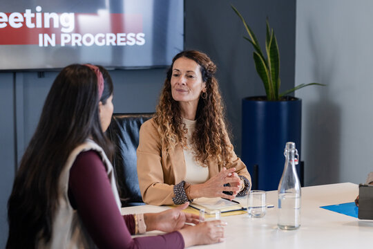 Diverse female colleagues in blazers discussing strategy at conference table with screen and notes