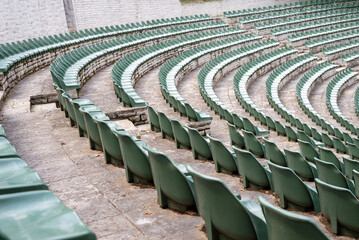 Large, open-air stone amphitheater is filled with numerous rows of green plastic seats arranged in...