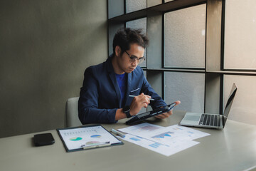 An Asian businessman sits tensely at his desk in a modern office, surrounded by documents and a laptop. He has a stressed expression, conveying a sense of pressure and concentration.