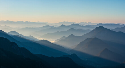 Aerial view of layered mountain ranges at dawn with soft blue sky and morning mist.
