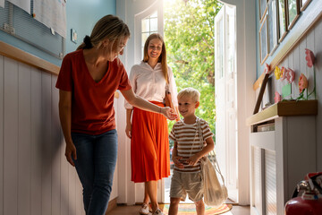 Mother saying goodbye to her son at preschool entrance as the teacher warmly guides him into the classroom.