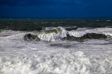 Powerful waves and churning ocean seen from the path to the Nazaré lighthouse in Portugal under a dramatic, moody sky.